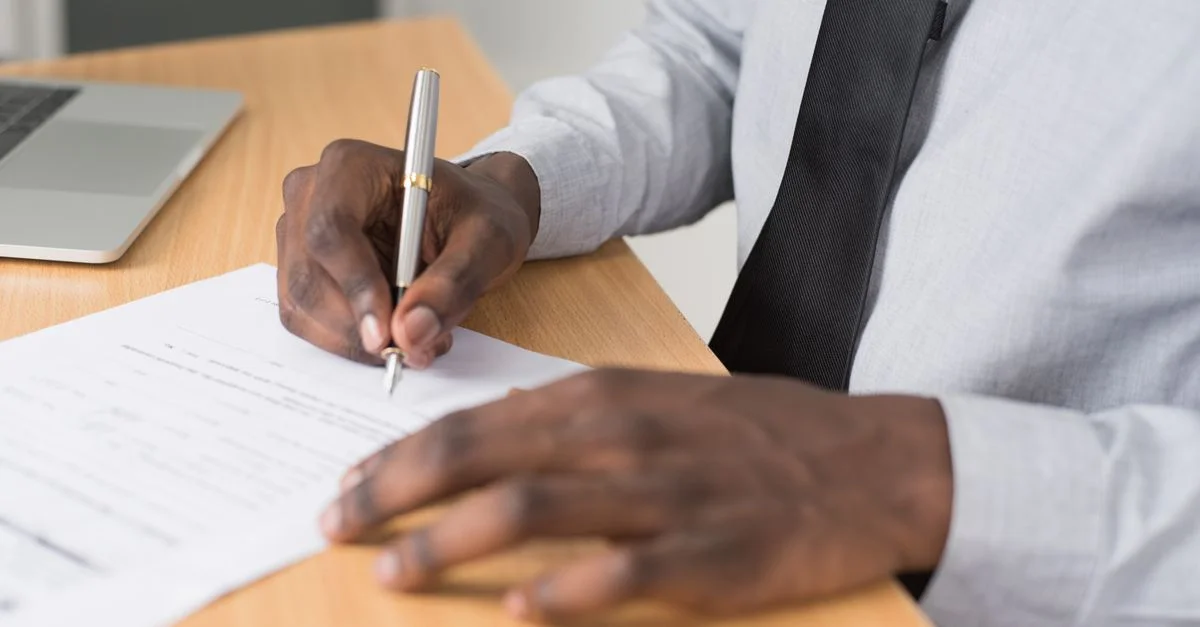 Gros plan sur un homme signant des documents sur un bureau, avec un ordinateur portable à proximité, dans un environnement de bureau professionnel.