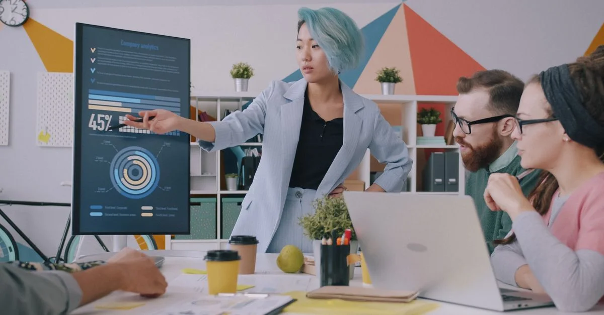 Réunion d'équipe diversifiée avec présentation de données à l'écran dans un environnement de bureau moderne.